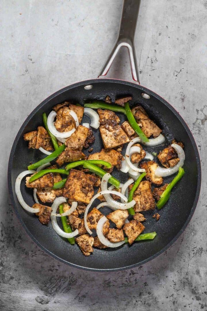 Marinated and baked tofu slices being sauteéd in a pan with cuts of onion and green peppers. 